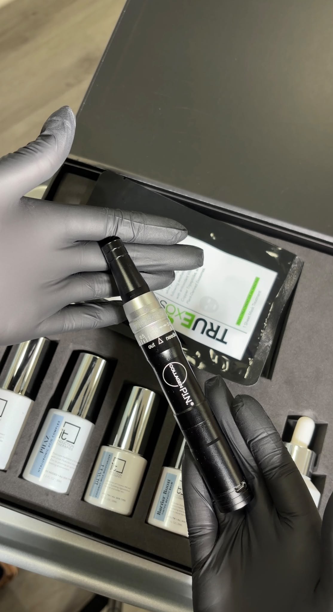 A gloved provider holding a Collagen P.I.N. microneedling pen above a kit of TrueExos and aftercare serums